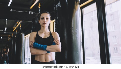 Serious smiling woman standing with crossed arms in the gym near the window, Sporty girl has wrapped her fists with blue cloth. Close up