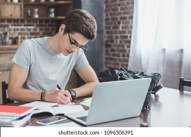 serious student writing homework at table with laptop and smartphone