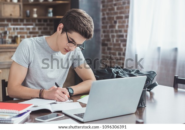 serious student writing homework at table with\
laptop and smartphone