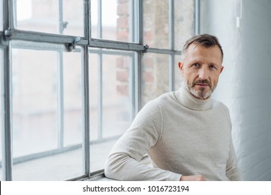 Serious Stylish Middle-aged Man Staring At The Camera With A Penetrating Look As He Leans Against The Sill Of A Large Window