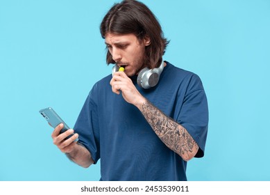 A serious tattooed Middle Eastern man in a T-shirt stands against a blue background, holding a phone and smoking a disposable electronic cigarette	