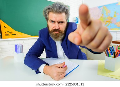 Serious teacher in classroom pointing finger. Bearded male sitting at table in class. Learning, education.