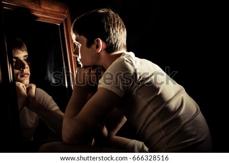 Serious teen boy surrounded by darkness looking at himself in mirror with wooden frame