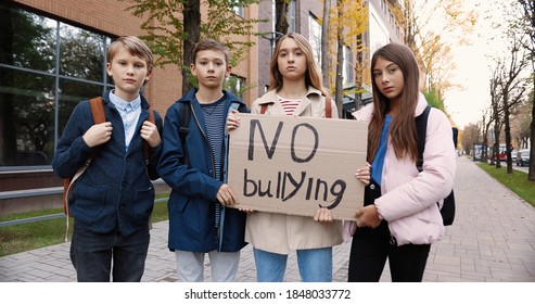 Serious Teen School Students Standing Outdoor Near School And Holding No Bullying Sign. Caucasian Boys And Girls Pupils With Poster Protesting Against Mocking And Intimidation. Social Issue Concept