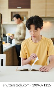 Serious Teenage Boy Reading Book When His Father Cooking Breakfast In Background