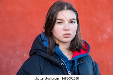 Serious Teenager Girl In A Jacket Stands Against A Red Wall Outdoors