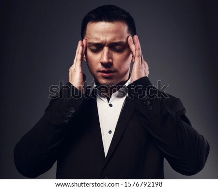 Serious thinking man with thought-transference abilities holding the hands the head with closed eyes in black suit on grey studio background. Closeup portrait. 