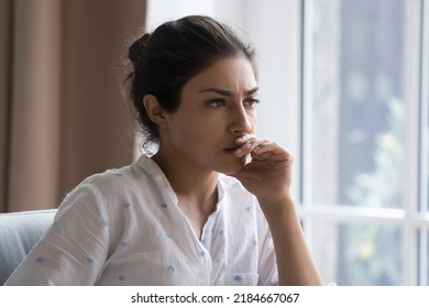 Serious thoughtful beautiful Indian woman sitting on couch indoors, touching chin, looking away, thinking over bad concerning news, problem, feeling worried, anxious, stressed, making decision