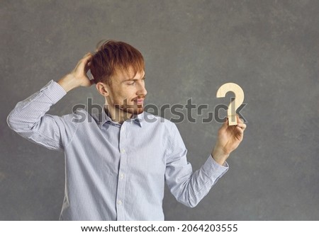 Serious thoughtful clever freelancer programmer man looking at wooden question figure in hand studio shot. Hipster red-haired young hipster showing puzzled confused undecided emotion scratching head