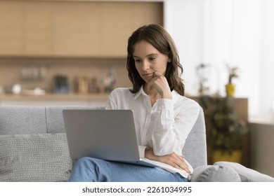 Serious thoughtful freelance employee girl using online app on laptop for job communication, working at computer, sitting on couch at home, reading email message, article, touching chin, thinking - Shutterstock ID 2261034995