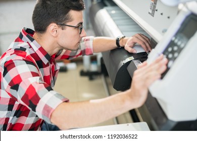 Serious thoughtful handsome young engineer in shirt examining wide format printer while holding inspection in printing house