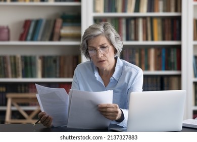 Serious Thoughtful Middle-aged Woman In Glasses Looks Worried Read News In Formal Document Sit At Workplace Desk With Wireless Computer. Older Female Review Paper Letter, Learns Report Feels Concerned