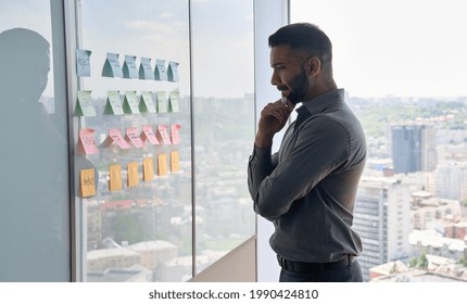 Serious Thoughtful Young Indian Businessman Ceo Near Whiteboard With Adhesive Sticky Notes Thinking About Ideas Of Planning Strategy Project Standing At Panoramic Window Of Modern Business Office.