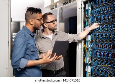 Serious Thoughtful Young Multi-ethnic Computer Support Specialists In Casual Clothing Standing At Server Racks And Using Laptop While Analyzing Network Problem