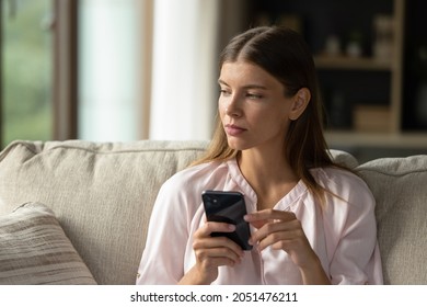 Serious thoughtful young woman holding smartphone, sitting on couch at home, thinking over online chat text message, email. Girl using mobile phone, getting bad news, problem, looking away - Powered by Shutterstock