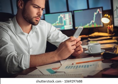 Serious Trader Using Mobile Phone App On Smartphone For Checking Stock Trading Data Analysis Concept, Focused Businessman Holding Cellphone Working In Office With Financial Graph On Computer Monitors