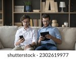 Serious twin brother working on family startup from home, doing freelance job, using mobile phone, tablet computer for online business communication, sitting on home couch together