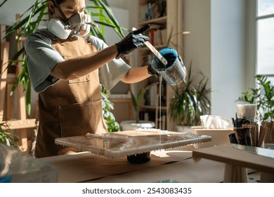 Serious woman artist works with epoxy resin, creating picture in technique of liquid art. Professional female wielding mixing stick, carefully pouring fluid on to canvas. Lady fills artwork with resin - Powered by Shutterstock