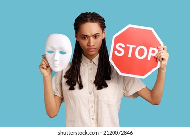 Serious Woman With Black Dreadlocks Holding White Mask And Red Stop Sign, Looking At Camera, Do Not Change Personality, Wearing White Shirt. Indoor Studio Shot Isolated On Blue Background.