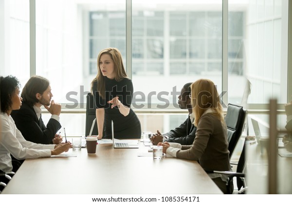 Serious woman boss scolding employees for bad\
results or discussing important instructions at multiracial team\
meeting, dissatisfied female executive talking to multiracial team\
at boardroom briefing