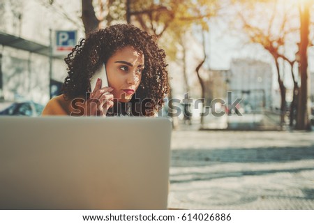 Serious woman calling with cell telephone while sitting in street cafe, attractive brunette curly lady having talking on smart phone while work on her laptop with copy space for your message or logo