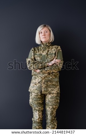 serious woman in camouflage standing with crossed arms isolated on black
