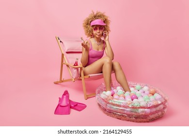 Serious Woman With Curly Hair Has Telephone Conversation Sits On Deck Chair Wears Swimsuit And Sunglasses Sits On Lounge Chair Stays At Home During Summer Vacation During Coronavirus Pandemic