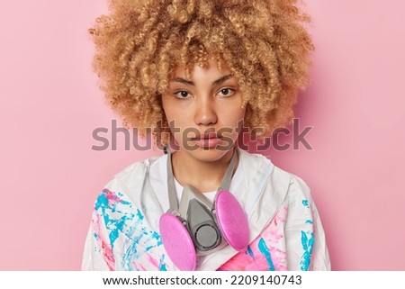 Serious woman with curly hair wears dirty protective suit and gasmask around neck focused directly at camera poses against pink studio background. People biohazard and biotechnology concept.