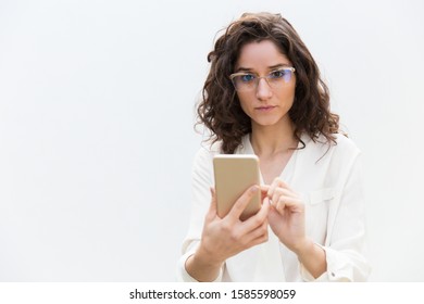 Serious woman in glasses using mobile app, texting message, consulting internet. Wavy haired young woman in casual shirt standing isolated over white background. Smartphone using concept