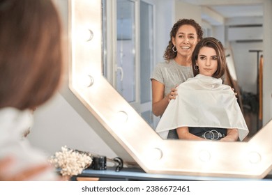 Serious woman in hairdresser cape on shoulders looking in mirror while stylist preparing hair in salon - Powered by Shutterstock