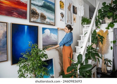 Serious woman hangs pictures on wall. Concentrated unsmiling female artist creates composition of paintings, decorations on wall. Creative designer in art home studio workshop among artworks plants. - Powered by Shutterstock