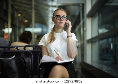 Serious Woman Intelligent Government Worker In Formal Wear And Spectacles Talking Via Mobile Phone And Using Notepad For Notary While Sitting In Airport Terminal Before Business Trip Abroad. 