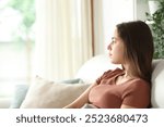 Serious woman looking through a window sitting on a sofa at home