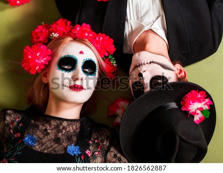Serious woman and man have traditional mexican image, wear sugar skulls, dressed in special attire for costume party, lie on the floor, isolated over green background. Female skull and male zombie