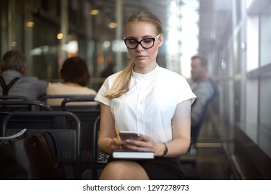 Serious Woman Office Worker Checking E-mail On Mobile Phone While Sitting In Waiting Room. Female Manager In Glasses Using Apps On Cell Telephone, Sitting In Airport Terminal Before Business Trip 