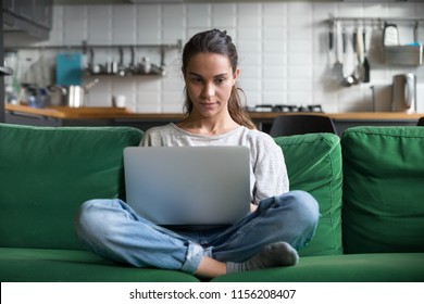 Serious woman using laptop checking email news online sitting on sofa, searching for friends in internet social networks or working on computer, writing blog or watching webinar, studying at home