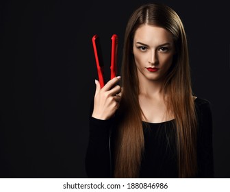 Serious woman young professional stylist with silky long hair wearing black clothes stands holding red hair straightener in hand at copy space over dark background. Haircare, beauty, wellness concept