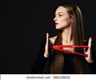 Serious woman young skillful professional stylist with silky long hair wearing black clothes stands holding red hair straightener looking aside at copy space. Haircare, beauty, wellness concept