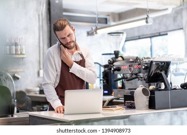 Serious Worried Barista Man Taking A Call On His Mobile Phone Checking Information Received On His Laptop With A Serious Concerned Expression In Cafe Shop. Startup Owner Small Business Concept.