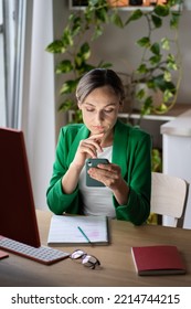 Serious Worried Woman Sitting At Workplace Desk Use Cellphone Read Message, Web Surfing. Focused Young Female Freelancer Scrolls Social Media And Reads News About Business And Projects In Internet. 