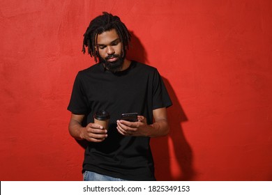 Serious Young African American Man With Dreadlocks 20s In Black Casual T-shirt Posing Hold Using Mobile Cell Phone Hold Paper Cup Of Coffee Or Tea Isolated On Red Color Background Studio Portrait