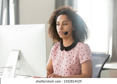 Serious young african call center operator customer care support manager in wireless headset talking using computer, mixed race telemarketer sales woman consulting client service helpdesk in office