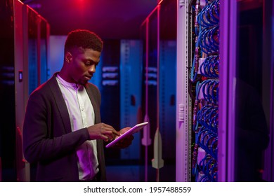 Serious young African-American data engineer standing at open server rack cabinet and using tablet while setting up system at data center - Powered by Shutterstock