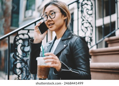 Serious young asian female in eyewear checking banking account during mobile conversation with operator holding ice coffee to go, young chinese  girl sitting on stairs talking on phone with friend - Powered by Shutterstock