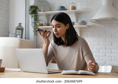 Serious young Asian student woman recording, sending audio message on smartphone, using voice recognition app. Remote employee working at laptop from home giving command to virtual assistant - Powered by Shutterstock