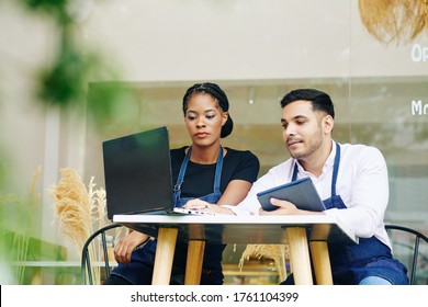 Serious Young Bakery Owners Sitting At Small Table, Discussing Financial Issues, Paying Bills And Ordering Products Online