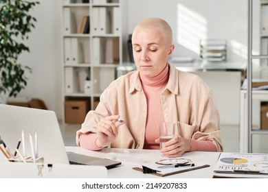 Serious Young Bald Woman In Turtleneck Sitting At Desk With Laptop And Holding Glass Of Water While Taking Pill In Office