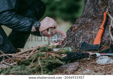 Serious young bearded hiker in cap setting fire in the forest using a flint rock. Various methods of starting a campfire. Survival skills.
