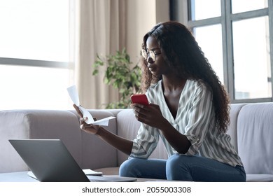 Serious Young Black African Woman Holding Paper Document Calculating Rent Or Money Savings, Paying Bills In Mobile Application On Cell Phone Doing Monthly Paperwork Sitting On Couch At Home.