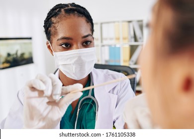 Serious young Black general practitioner in medical mask asking patient to open mouth so she could check her throat